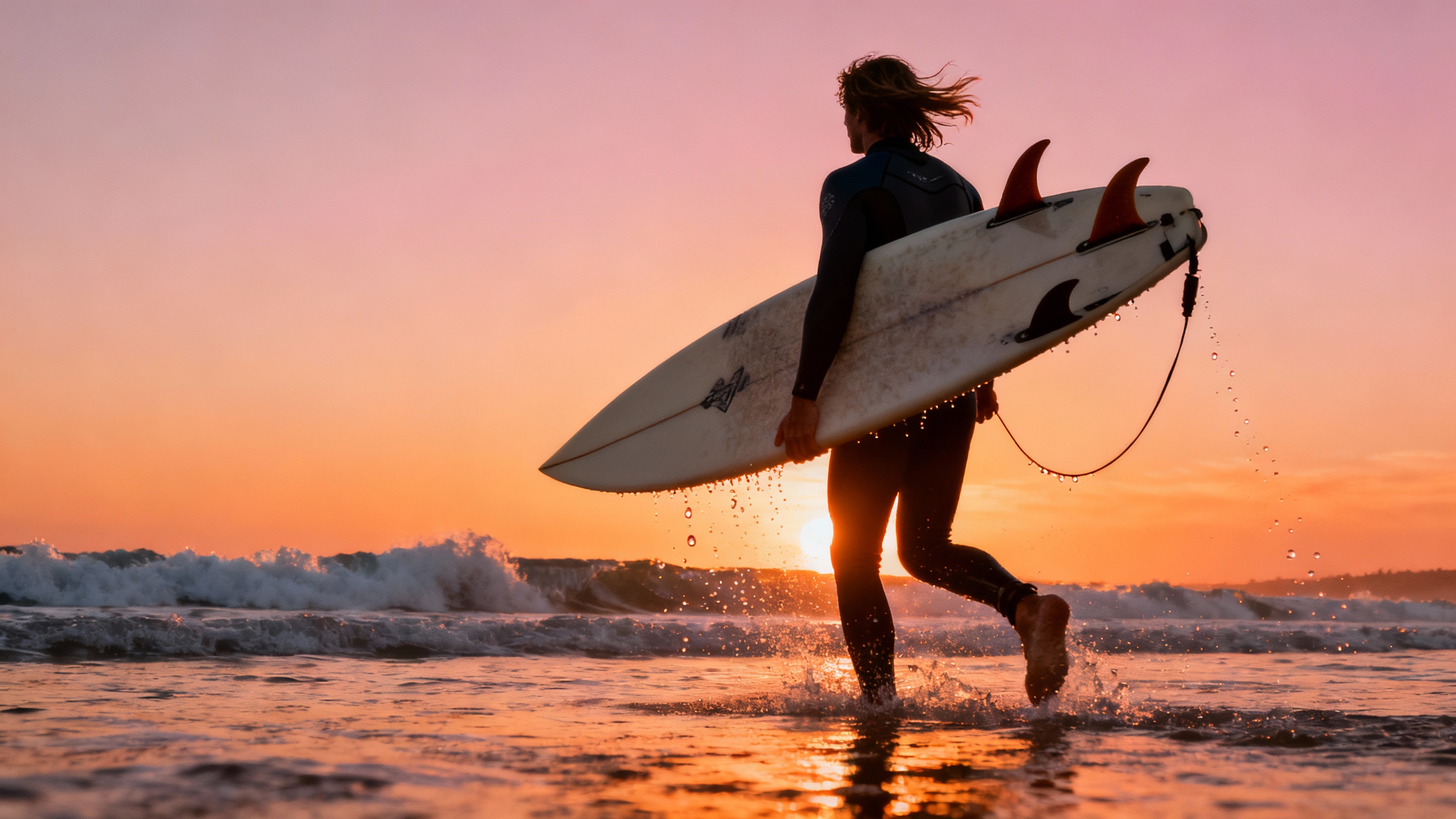 Dynamic action shot of a surfer carrying a surfboard under their arm, heading toward the ocean at sunrise, waves crashing dramatically in the distance.