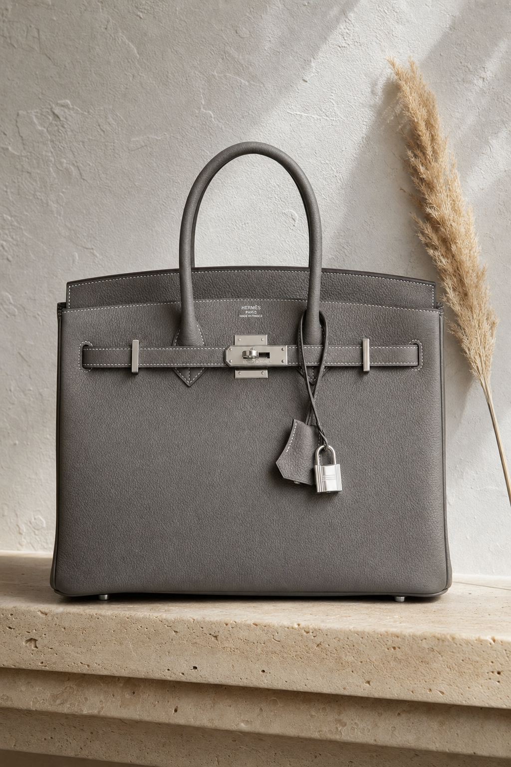 A tall 9:16 vertical luxury product photograph of a Hermès Birkin 30 in Togo leather, Etain gray colorway, placed upright on a smooth cream travertine stone shelf mounted against a raw white plaster wall. The structured bag stands perfectly centered in the frame, its palladium hardware — turn-lock, clochette, and lock — each catching a soft directional light from the upper left at slightly different angles, creating individual highlights on every metal element. The iconic double stitching along every seam is visible at full zoom. The Hermès Paris stamp on the front hardware is clearly legible. A single dried pampas grass stem leans casually against the right side of the bag. Shot on an 85mm lens at f/4. Color grade: warm travertine stone, cool plaster wall, the gray leather as the tonal hero. Mood: old money, architectural, gallery-level luxury product photography.
