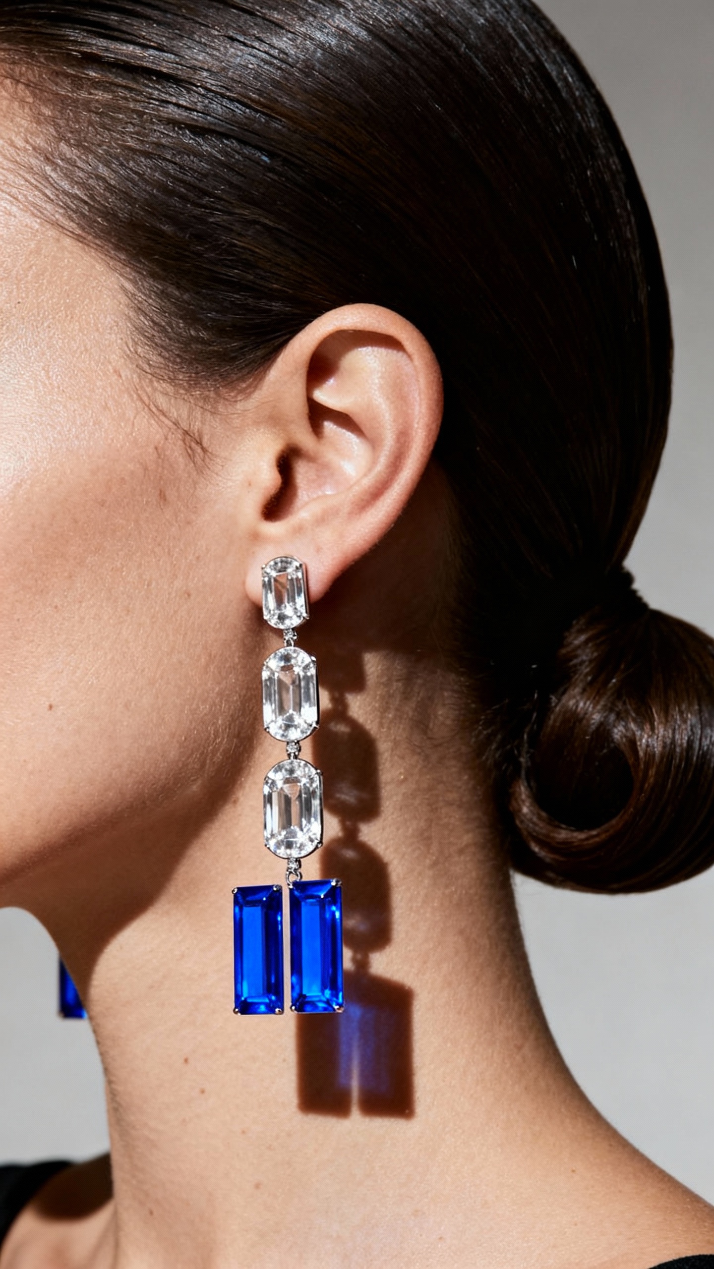 a striking close-up portrait showcasing a woman’s ear adorned with an elegant pair of long, luxurious earrings. The earrings feature a geometric design made up of rectangular and oval-cut clear gemstones that cascade downward, ending with two vivid blue rectangular gemstones that add a bold pop of color.

The lighting is dramatic and directional, highlighting the texture and sheen of the skin as well as the brilliant sparkle and clarity of the gemstones. The subject’s hair is sleeked back tightly, creating a clean and sculptural look that draws full attention to the jewelry. The overall aesthetic is sophisticated and high-fashion, exuding modern glamour and refined luxury.